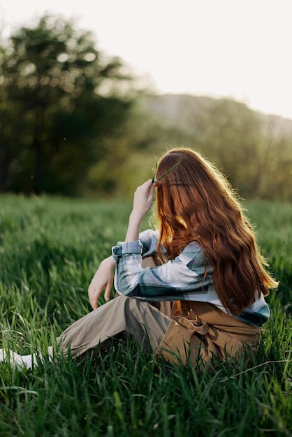 Kobieta siedzi na zielonej trawie w parku plecami do aparatu i relaksuje się na łonie natury w letnim świetle wieczornego zachodu słońca