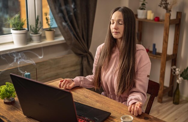Kobieta siedzi przy stole przed laptopem, medytując i relaksując się, słuchając instruktora online lub wykładu wideo Koncepcja zdalnej terapii i sesji medytacyjnej
