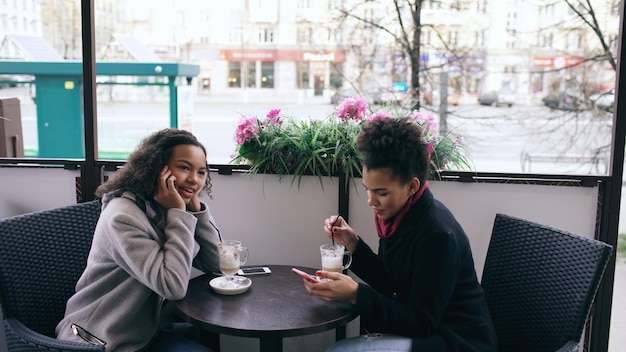 kobieta siedzi przy stole w ulicznej kawiarni rozmawiając przez telefon komórkowy, podczas gdy jej przyjaciółka surfuje po smartfonie