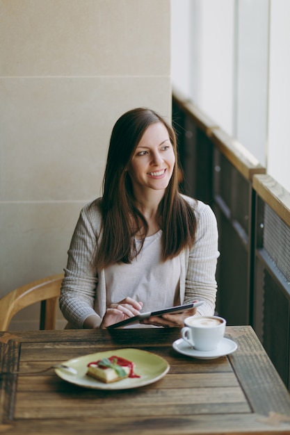 Kobieta siedzi sama w pobliżu duże okno w kawiarni przy stole z filiżanką cappuccino, ciasto, relaks w restauracji w czasie wolnym. Kobieta pracuje na komputerze typu tablet pc odpocząć w kawiarni. Koncepcja stylu życia.