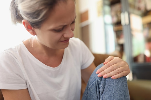 Zdjęcie kobieta siedzi w domu i czuje ból w kolanie nieszczęśliwa kobieta cierpiąca i dotykająca kolana