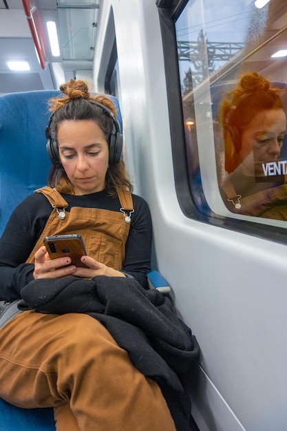 Kobieta siedzi w pociągu, patrzy na swój telefon i pisze SMS-a na słuchawkach.