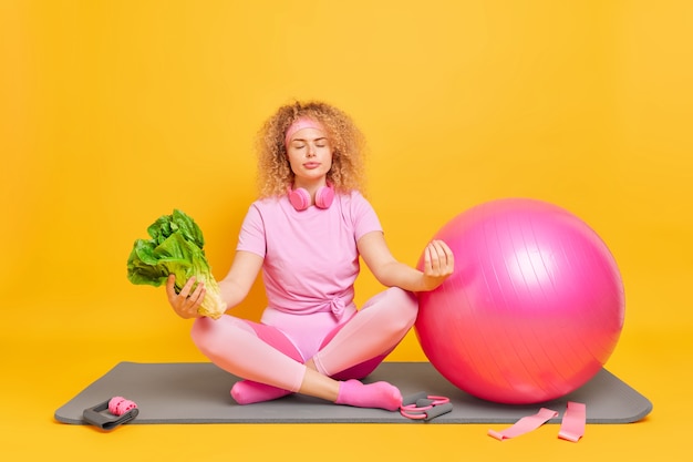 kobieta siedzi w pozycji lotosu próbuje się zrelaksować, medytuje po treningu fitness, ćwiczenia jogi trzyma zielone warzywo na macie w pobliżu sprzętu sportowego fitball