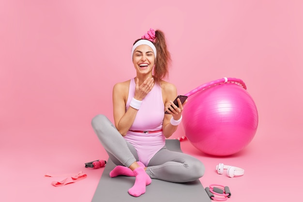 kobieta śmieje się z radością trzyma telefon komórkowy cieszy się treningiem fitness ubrana w body siedzi na macie otoczona fitballem z hula-hoop