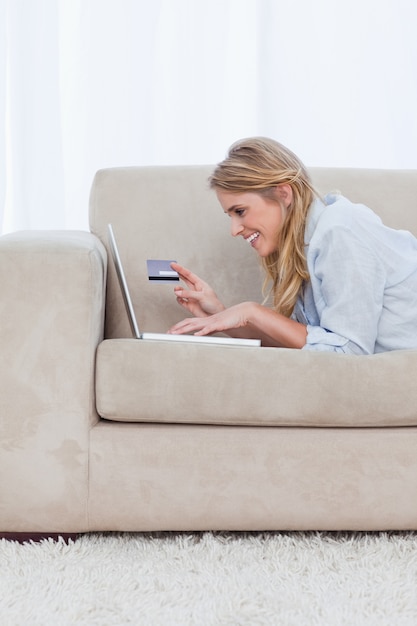 Kobieta spoczywająca na łokciach trzyma kartę bankową i pisząc na swoim laptopie