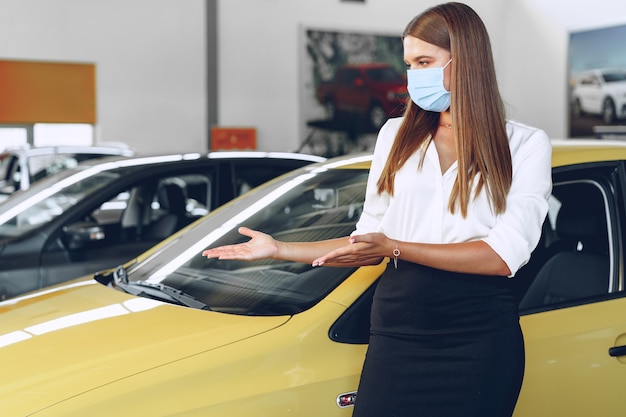 Kobieta sprzedawca samochodów stojących w pobliżu nowego samochodu na sobie ochronną maskę na twarz w salonie