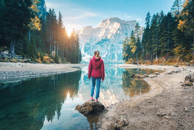 Kobieta stoi na kamieniu nad jeziorem Braies o wschodzie słońca jesienią Dolomity Włochy Krajobraz z dziewczyną górskie jezioro piękne odbicie w wodzie drzewa niebo ze słońcem Las jesienią Podróż