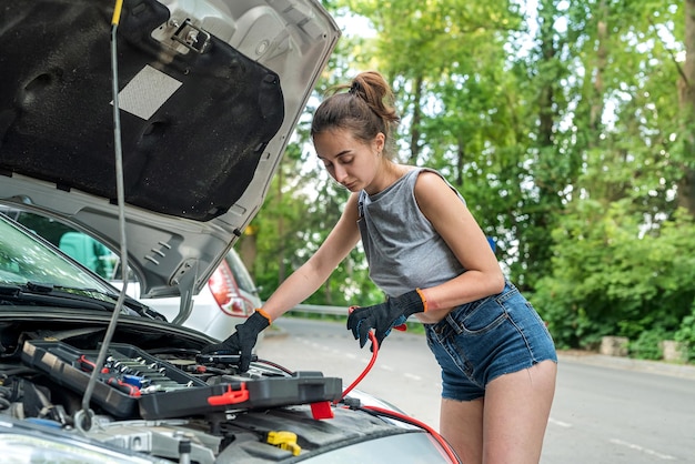 Kobieta Stoi W Pobliżu Zepsutego Samochodu I Trzyma Czarno-czerwony Kabel Połączeniowy, Aby Naładować Akumulator Auto Naprawy Samochodu Podczas Letniej Wycieczki