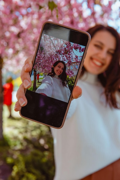 Kobieta strzelająca do siebie przez telefon pod kwitnącym drzewem sakura