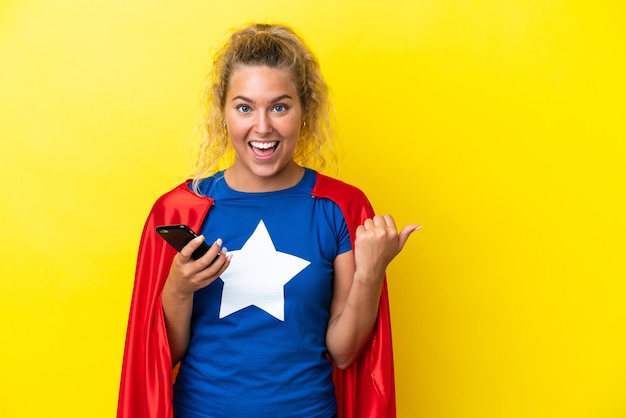 Kobieta Super Hero odizolowana na żółtym tle za pomocą telefonu komórkowego i wskazująca na bok