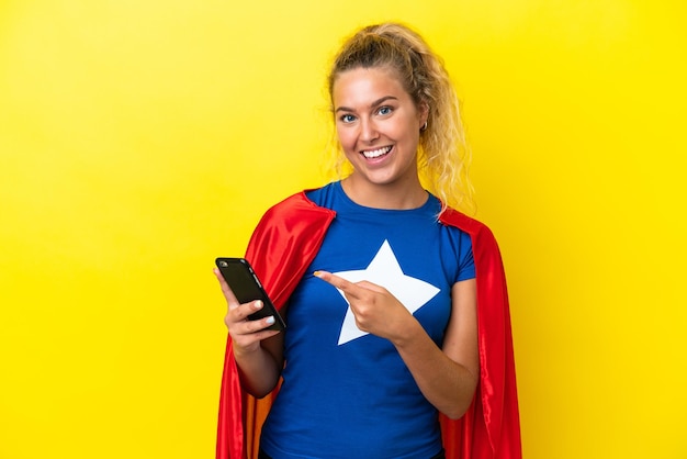 Kobieta superbohater na żółtym tle za pomocą telefonu komórkowego i wskazując go