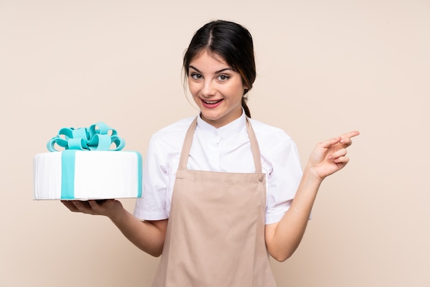Kobieta szefa kuchni ciasta trzymając wielki tort nad ścianą zaskoczony i wskazując palcem na bok