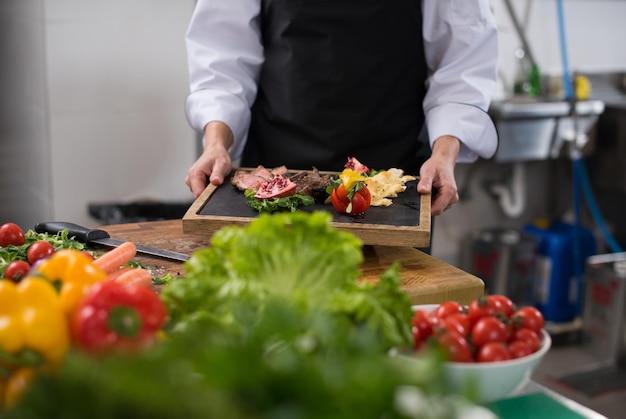 Kobieta szefa kuchni w kuchni hotelowej lub restauracyjnej trzymająca talerz z grillowanym stekiem wołowym z dekoracją warzywną