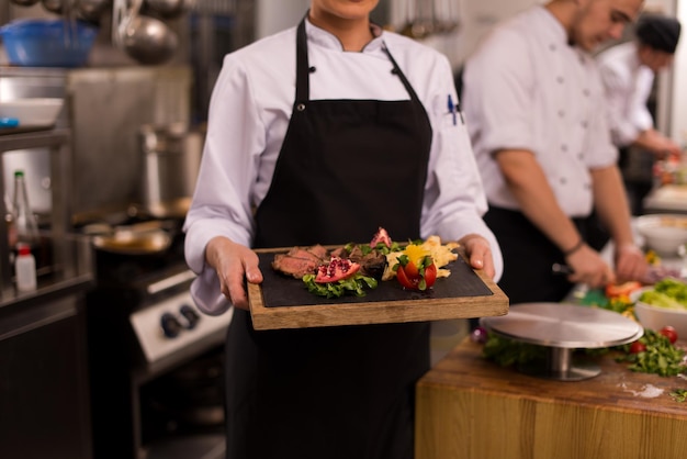Kobieta szefa kuchni w kuchni hotelowej lub restauracyjnej trzymająca talerz z grillowanym stekiem wołowym z dekoracją warzywną