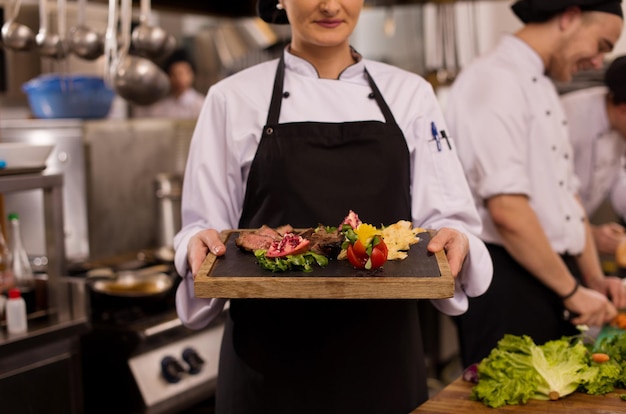 Kobieta szefa kuchni w kuchni hotelowej lub restauracyjnej trzymająca talerz z grillowanym stekiem wołowym z dekoracją warzywną