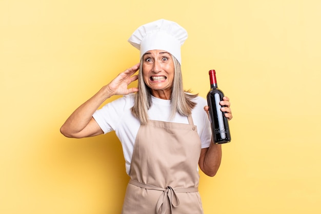 Kobieta szefowa kuchni w średnim wieku czuje się zestresowana, zmartwiona, niespokojna lub przestraszona, z rękami na głowie, panikując z powodu błędu, trzymając butelkę wina