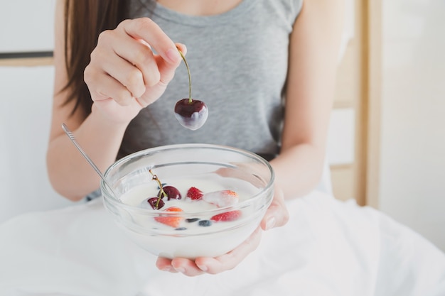 Zdjęcie kobieta trzyma szklaną miskę jogurtu i truskawek na białym łóżku rano.