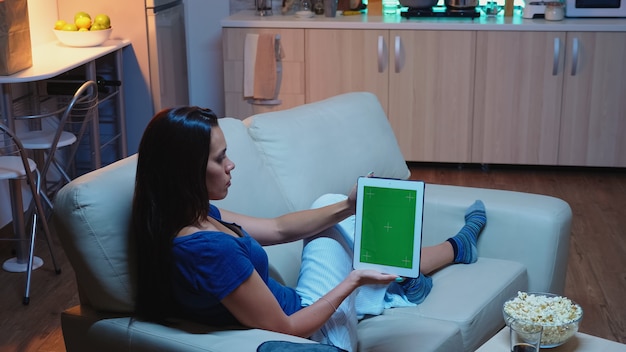 Kobieta trzyma touchpada z zielonym ekranem, siedząc na kanapie w salonie. Gospodyni domowa patrząc na szablon chroma klucz na białym tle inteligentny wyświetlacz notebooka przy użyciu technologii internet siedząc na kanapie.