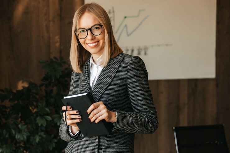 Zdjęcie kobieta trzyma w rękach notebook, stoi i uśmiecha się w biurze, planując nowe projekty dla swojej firmy.
