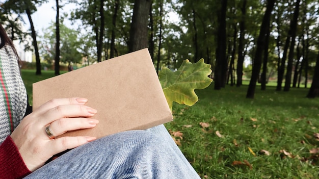 Kobieta trzyma zamkniętą książkę leżącą na kolanach z zbliżeniem opadłego liścia dębu w parku w słoneczny, ciepły jesienny dzień. Pojęcie samego relaksu, czytania i relaksu.