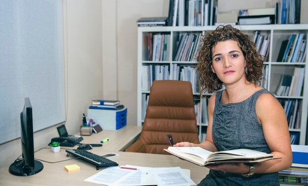 Kobieta trzymająca notatnik siedząca nad biurkiem w biurze i patrząca