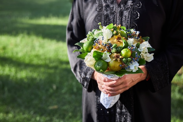 Kobieta ubrana w abaya stoi z bukietem kwiatów i owoców w dłoniach