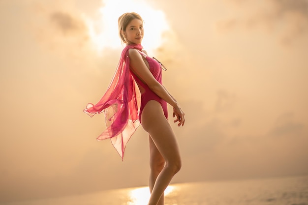 Kobieta ubrana w różowy jednoczęściowy strój kąpielowy, trzymająca szalik na wietrze, cieszyć się romantycznym momentem zachodu słońca na kamiennej tropikalnej plaży