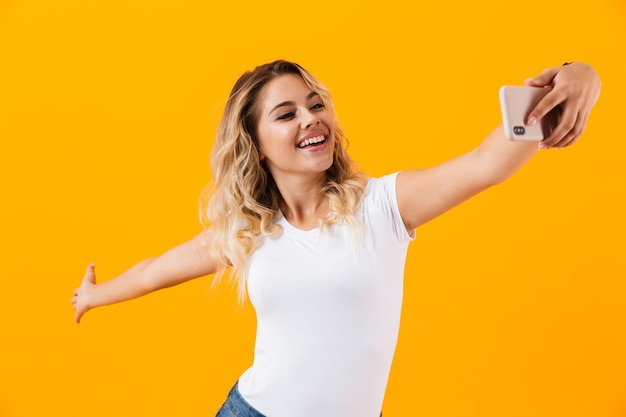Kobieta uśmiecha się i biorąc zdjęcie selfie na telefon komórkowy, na białym tle nad żółtą ścianą