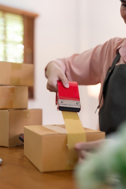 Kobieta używa taśmy do przymocowania pudełek do paczek, aby przygotować towary do procesu pakowania, wysyłki, sprzedaży online, marketingu internetowego, koncepcji e-commerce, początkującego pomysłu biznesowego