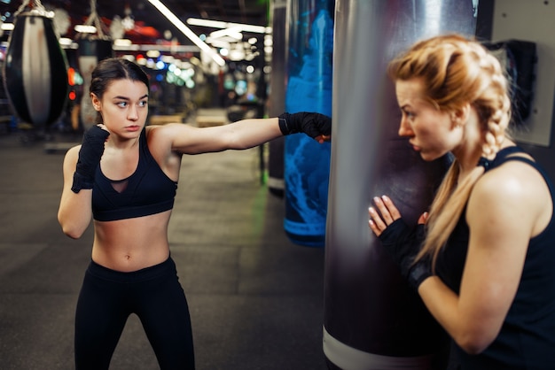 Kobieta W Bandażach Bokserskich Uderza W Worek Treningowy, Trenuje Boks Z Trenerem. Bokserki Na Siłowni, Trening Kickboxingu W Klubie Sportowym, ćwiczenie Ciosów Pięścią