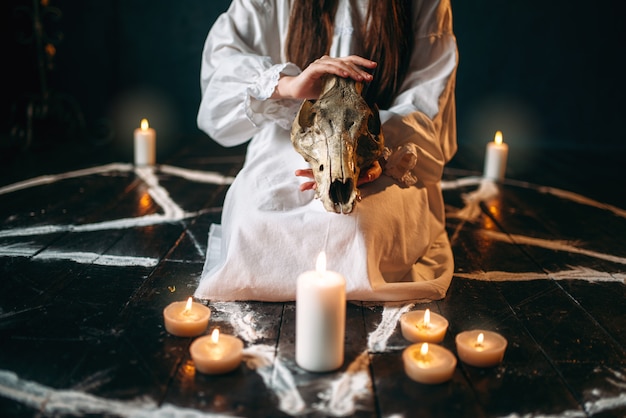 Zdjęcie kobieta w białej koszuli trzyma w rękach zwierzęcą czaszkę, koło pentagramu ze świecami. rytuał czarnej magii, okultyzm