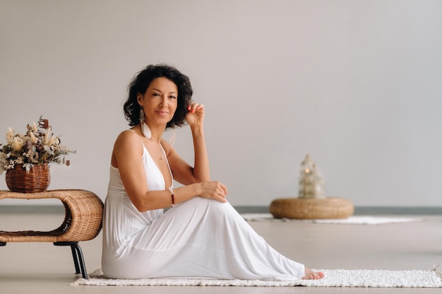 Kobieta w białej sukience w jasnym wnętrzu siedzi na białym dywaniku. Pojęcie relaksu.