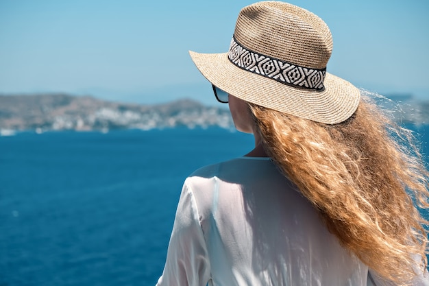 kobieta w białej sukni kąpielowej i słomkowym kapeluszu na białym tarasie balkon z widokiem na morze