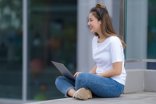 Kobieta w biały t-shirt i niebieskie dżinsy za pomocą laptopa siedząc na podłodze na zewnątrz w tle ulicy miasta, letni dzień