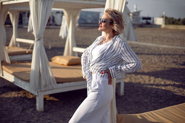 Zdjęcie kobieta w białych ubraniach i okularach przeciwsłonecznych stoi na plaży w pobliżu drewnianego