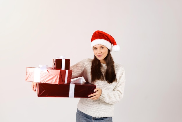 Zdjęcie kobieta w białym swetrze trzymająca pudełka świąteczne z prezentem w kapeluszu św. mikołaja izolowana na szarym tle