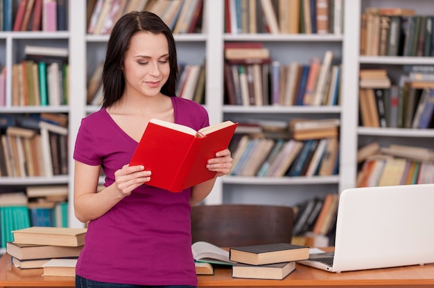 Kobieta w bibliotece. Wesoła młoda kobieta czyta książkę, opierając się przy biurku w bibliotece