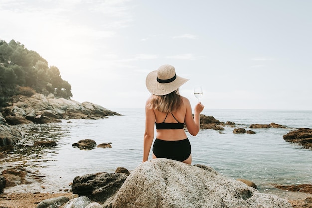 Kobieta w bikini i kapeluszu sama na plaży trzymająca kieliszek wina i ciesząca się spokojną scenerią widok z tyłu Kobieta ciesząca się czasem spędzonym na plaży śródziemnomorskiej opalając się i pijąc białe wino