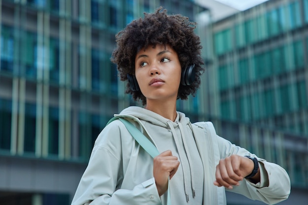 kobieta w bluzie sprawdza wyniki fitness na smartwatchu odwraca wzrok słucha muzyki przez słuchawki pozuje na tle nowoczesnego szklanego budynku