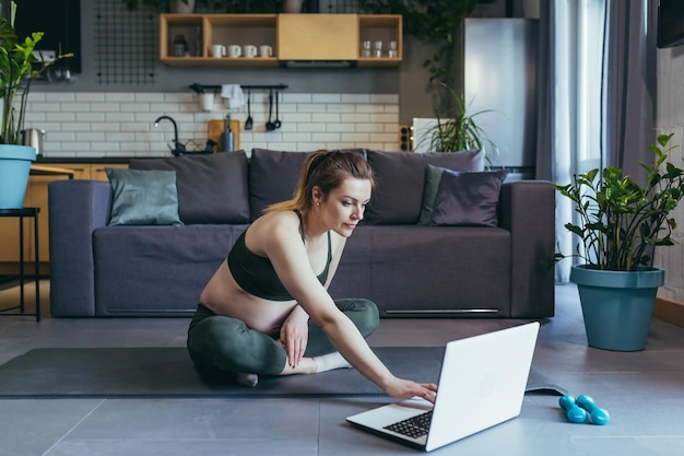 Kobieta w ciąży ćwicząca fitness w domu używa laptopa do zajęć online