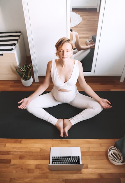 Kobieta w ciąży ćwiczy jogę w domu z laptopem Ćwiczenia kobiece medytują podczas ciąży