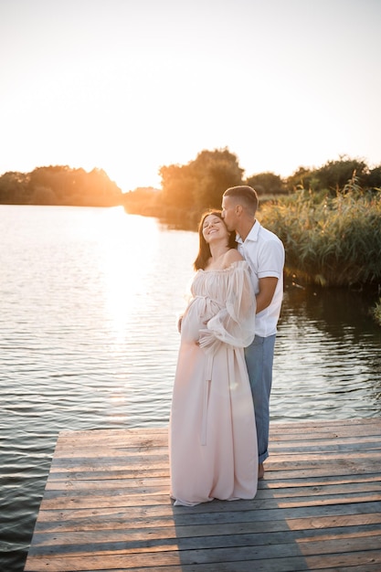 Kobieta w ciąży i jej mąż stoją na molo i patrzą na siebie piękna rzeka i zachód słońca przyszli rodzice spędzają czas na świeżym powietrzu piękna młoda matka