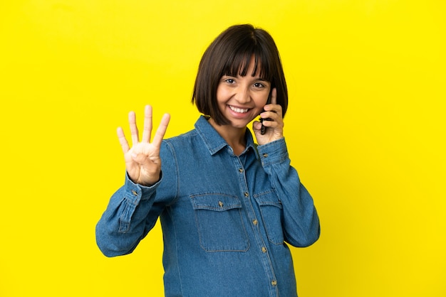 Kobieta w ciąży korzystająca z telefonu komórkowego na żółtym tle szczęśliwa i licząca cztery palcami