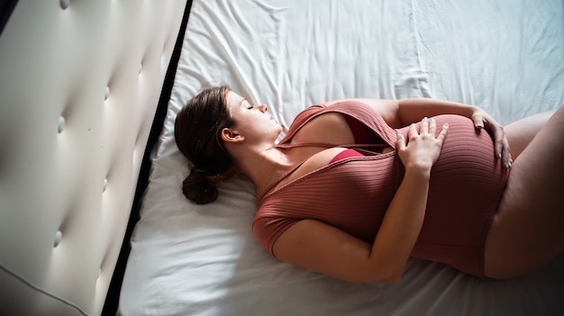 Kobieta w ciąży leży na białym łóżku i prasuje brzuch. Widok z góry