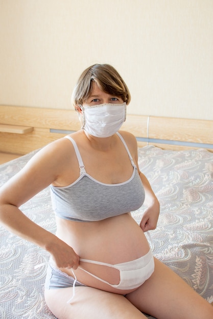 Kobieta W Ciąży Maska Medyczna I Maska Na Brzuchu W Ciąży, Siedzi Na łóżku.