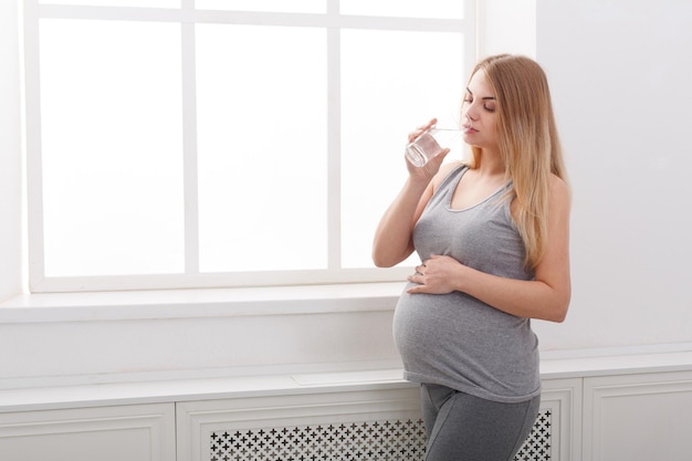 Kobieta w ciąży pije szklankę mleka kopii przestrzeni. Młoda blondynka oczekująca, ciesząc się zdrowym napojem, stojąc w pobliżu okna. Opieka zdrowotna, odżywianie, witaminy, koncepcja ciąży