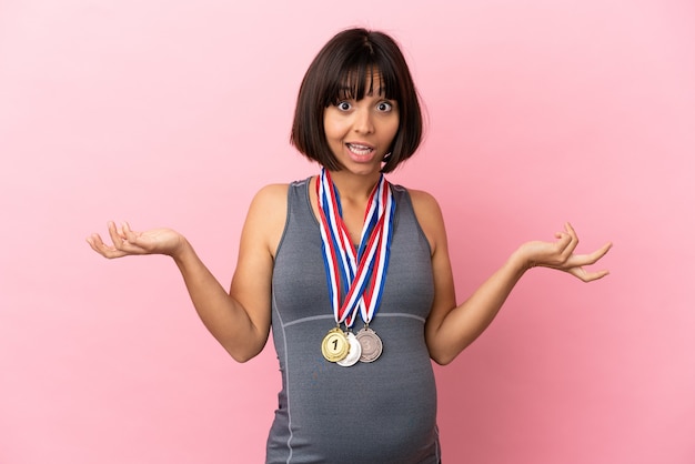 Zdjęcie kobieta w ciąży rasy mieszanej z medalami na różowym tle ze zszokowanym wyrazem twarzy