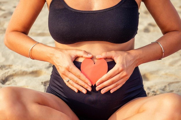 Kobieta w ciąży robi joga na plaży trzymając czerwoną kartkę papieru w kształcie serca i maty do jogi.