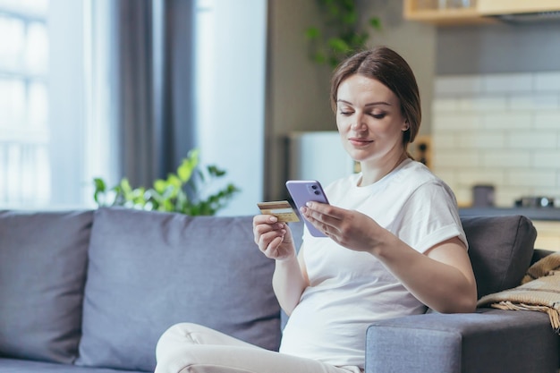 Kobieta w ciąży siedzi na kanapie w domu, używając telefonu podczas zakupów online, trzymając bankową kartę kredytową