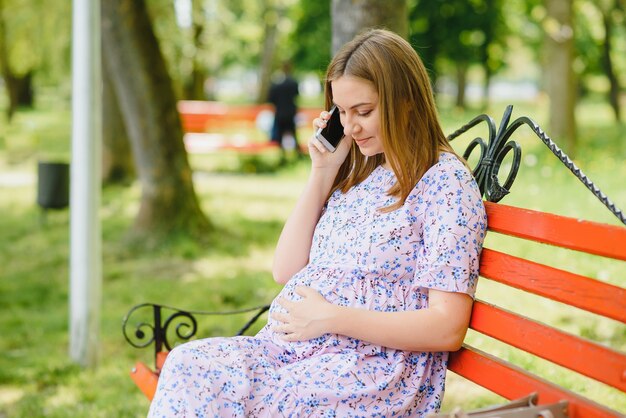 Kobieta w ciąży siedzi na ławce w parku i przy użyciu telefonu